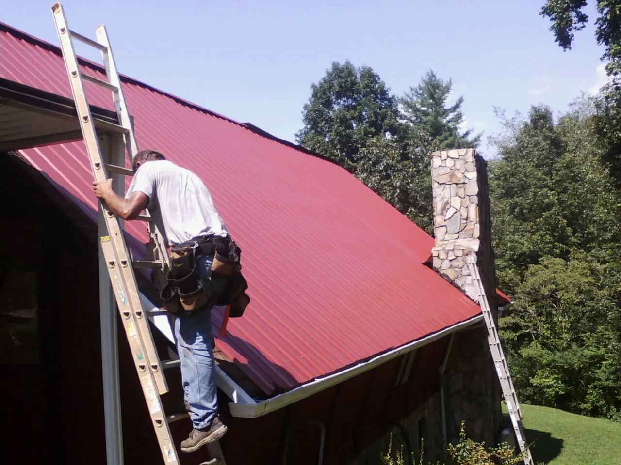 Roofer on a ladder beside a red metal roof and stone chimney — Soffit & Fascia Repair by Roofing Brothers in O Brien